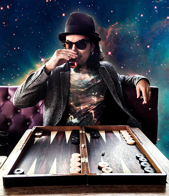Photo of Memo Akten in hat and sunglasses, sitting at a booth behind a backgammon board, with outerspace background