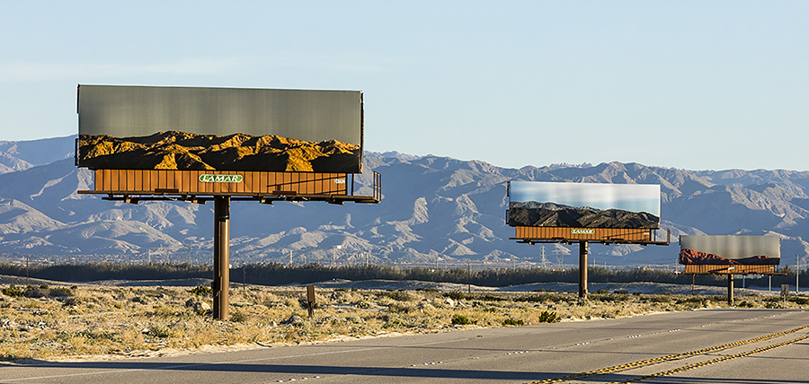 Photograph of a road with mountains in the distance. The road is lined with three large billboards depicting photos of those same mountains at different times of day.
