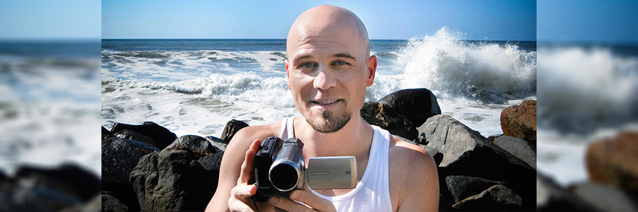 Photo of Adam Kamil holding a video camera with the ocean behind him.