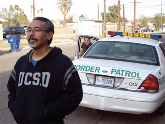 photo of Ricardo Dominguez with a serene smile, wearing a UCSD sweatshirt, as border patrol approach him from behind