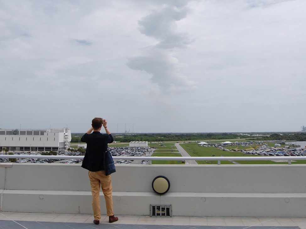 Photo from behind Sascha Pohflepp as he looks out over a large parking lot and field with large gray sky above