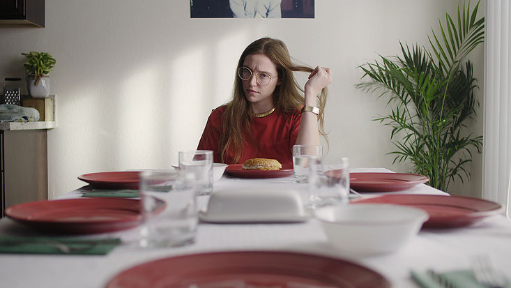 Photo of Wren Gardiner sitting at the head of a dining table set with empty red plates. Only her plate has a bagel. has 