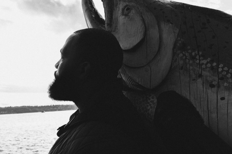Photo of Amir Saadiq facing away from camera, leaning against a large wooden fish, looking out over water